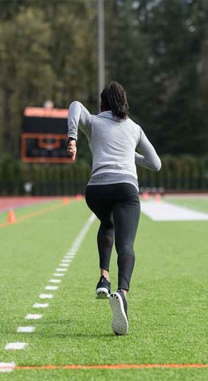 An African American female track athlete trains at a stadium. She is running sprints on the field. The shot is from behind her as she runs away from frame.